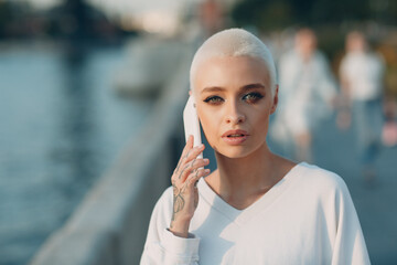 Wall Mural - Portrait of young smiling millenial european short haired woman using mobile phone. Beautiful happy blonde girl speaking cellphone outdoor. Summer fashion female clothing.