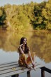 © SHOTPRIME STUDIO - A young happy woman with a hippie smile sits on the lake shore on a bridge wearing eco clothing made of natural materials in harmony with nature in the fall. Travel lifestyle