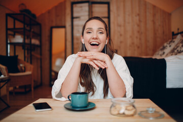 Smiling happy young woman sitting at table at home behind computer laptop and talking on video call. Girl female with cup of tea or coffee speaking online on webcam indoors