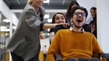 Cheerful People Have Fun During Workday Break. Two Teams Compete In Chair On Wheels Racing In Office Hallway. Colleagues Pushing Coworkers On Chairs. Winners Rejoice. Employees Film On Smartphones.