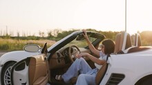 Beautiful Stylish Woman Goes To Her Luxury White Convertible. Back View. Female Sits Down In The Car And Close Door. Sun Sets Background.