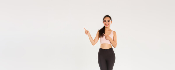 Wall Mural - Full length of smiling, cute slim asian girl in sportswear, pointing fingers upper right corner, inviting to buy gym membership, showing workout offer, fitness coach show healthy diet recommendations