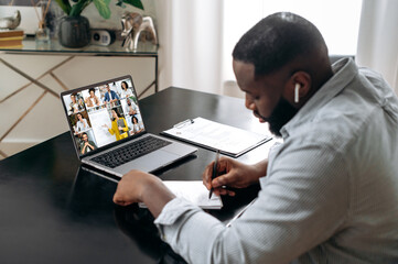 Wall Mural - Online training, lecture, seminar. African american smart student listening to the online lecture, taking notes in notebook, on the laptop screen are teacher and the group of multiracial students