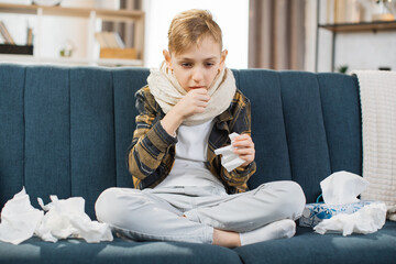 Pleasant tired sick teen boy with scarf around neck, sitting on soft couch and coughing, suffering from flu or cold and sore throat. Flu treatment at home.