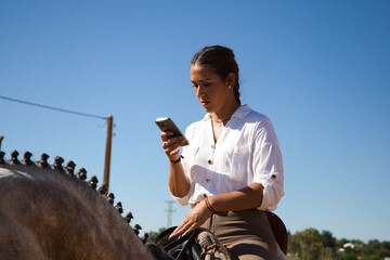 Young and beautiful Spanish woman on a white and grey horse consulting her mobile phone. The woman is wearing a riding uniform. Thoroughbred and equine concept.