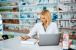© Drazen - Female pharmacist going through paperwork while working on laptop in pharmacy.