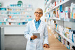 © Drazen - Happy senior pharmacist using digital tablet in pharmacy and looking at camera.