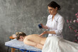 Portrait of serious middle-aged masseuse pouring massage aroma oil from blue plastic cup on bare back of woman patient.
