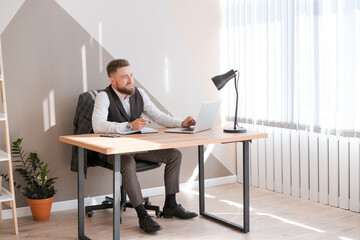 Caucasian bearded business man is sitting at window table using his laptop and writing notes in a notebook while doing office work. In a business suit doing his job