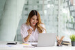 © amnaj - Smiling beautiful Asian businesswoman sitting happily and comfortably working on her laptop computer and taking notes and working on paperwork at the office.