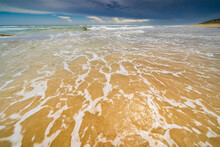 Low Wide Angle View Of Waves Rolling Up A Golden Sandy Beach With Dark Storm Clouds Overhead
