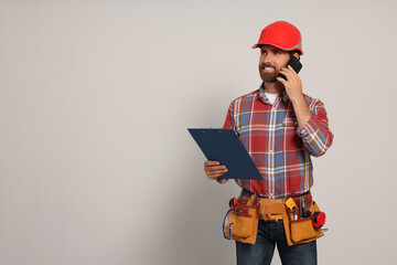 Poster - Professional builder in hard hat with clipboard and tool belt talking on phone against light background, space for text