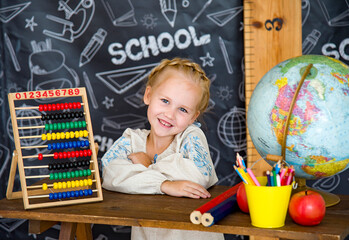 little smiling blonde girl in linen embroidery sitting at the desk near blackboard with abacus,  globe and colored pencils  in classroom at school