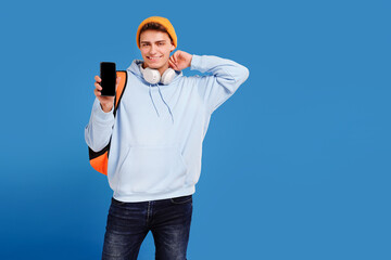 Photo of young fashionable student guy with cap and backpack smiling at camera, showing empty screen of mobile phone, isolated over blue studio background.