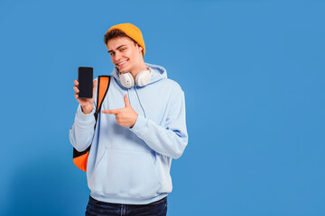 Photo of young fashionable student guy with cap and backpack smiling at camera, pointing on empty screen of mobile phone, isolated over blue studio background