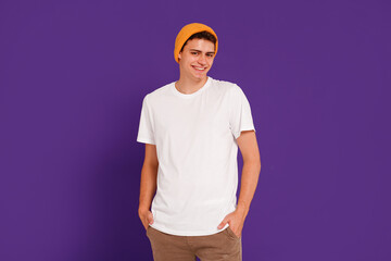 Wall Mural - Portrait of happy attractive hipster boy in good mood posing in yellow cap, smiling to the camera, standing on violet studio background.