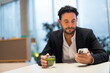 Portrait of happy handsome Hispanic businessman at coffee shop having green tea while using phone