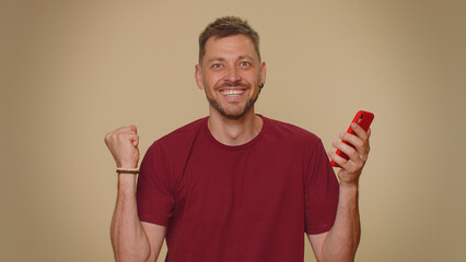 Happy excited joyful young man use smartphone typing browsing say wow yes, found out great big win news doing winner gesture. Adult stylish male guy isolated on beige studio wall background indoors