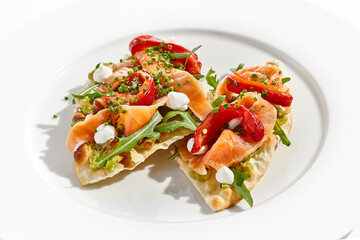 Canvas Print - Bruschetta with salmon, avocado and paprika on white plate. Salmon bruschetta on crispy focaccia. Appetizer with guacamole, trout, cheese and paprika on toast. Fish antipasti in restaurant menu.