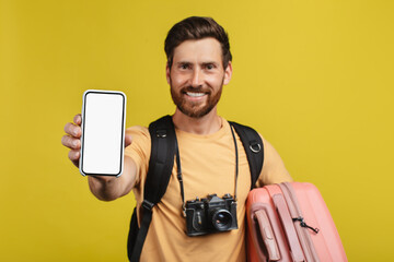 Happy man showing smartphone empty screen, wearing backpack and camera, standing with suitcase, mockup