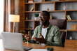 © Prostock-studio - Laughing middle aged african american male in glasses works on laptop at table, have meeting, gesticulate