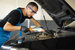 © uflypro - The mechanic in blue overalls and safety glasses inspects the car while working with led lamp. The work of a car mechanic in a modern workshop, car service.
