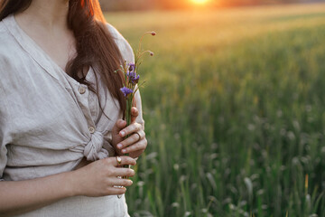 Wall Mural - Woman holding wildflowers in wheat field in warm sunset light. Close up of young female in rustic dress with flowers in hands in evening summer countryside. Tranquil atmospheric moment