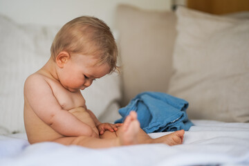 cute naked one year old happy blond baby boy sitting at home on a cozy bed after bathing and looking
