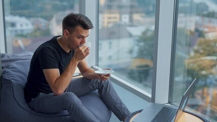 Wall Mural - Brunet young man drinking coffee at lunch break sitting near panoramic window. Thoughtful male sitting in front of laptop. Blurred backdrop.