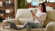© DC Studio - Beautiful woman having smartphone while working remotely and sitting on sofa at home. Attractive person with touchscreen modern device enjoying remote work while relaxing on couch in living room.