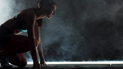 Wall Mural - Silhouette female runner in race  start position. Girl in sportswear posing on lit track with smoke in the background.