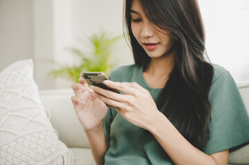 Wall Mural - Happy young asian woman relaxing at home she is sitting on sofa and using mobile smartphone