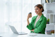 © amnaj - Pretty Asian businesswoman sitting happily working on her laptop and drinking coffee smiling and laughing brightly in the office.