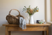 Wicker Basket With Grey Scarf, Australian Native Flowers In White Jug And Assorted Bowls On Oak Side Table Next To Window
