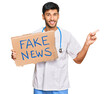 Young handsome man wearing doctor uniform holding fake news banner smiling happy pointing with hand and finger to the side