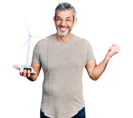 Wall Mural - Middle age hispanic with grey hair holding solar windmill for renewable electricity screaming proud, celebrating victory and success very excited with raised arm