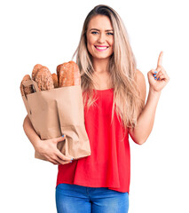 Sticker - Young beautiful blonde woman holding paper bag with bread surprised with an idea or question pointing finger with happy face, number one