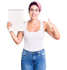 Sticker - Young beautiful woman with pink hair holding notebook smiling happy and positive, thumb up doing excellent and approval sign