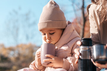 A cute little girl is drinking tea with her mom at a picnic in an autumn park on a sunny day.Family and autumn concept.Selective focus,copy space.