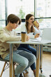 © LIGHTFIELD STUDIOS - teenage girl drinking coffee near laptop and friend writing in library reading room.