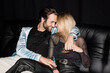 © LIGHTFIELD STUDIOS - Positive man in jacket hugging blonde girlfriend on leather couch isolated on black.