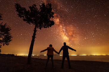 Wall Mural - Two hikers silhouette stands on the shore on the milky way galaxy background. Dark starry night.
