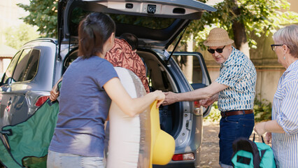 Wall Mural - Big family packing voyage luggage in car trunk while getting ready for summer field trip. Happy people loading vehicle with luggage and trolleys while going on weekend citybreak together.