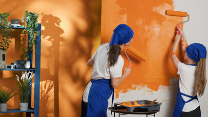 Wall Mural - People painting walls with orange color and roller brush, using tools and equipment to paint home interior decor. Mother and young child having fun redecorating apartment room. Tripod shot.