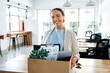 Happy young brazilian or hispanic female employee, holding cardboard box, standing in modern office and looking at camera, smiling, newcomer woman got new job, her first day of work in new workspace