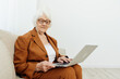© Tatiana - a joyful, sweet, gray-haired elderly woman in a brown suit is sitting on a beige sofa in a bright room working remotely on a laptop
