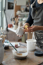Female Artisan Enjoys Process Of Handmade Pottery Craft Standing At Round Wooden Table With Painting Equipment. Woman In Black Apron Creates White Ceramic Mug On Blurred Background In Workshop Closeup