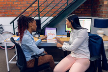 Multiracial Young Businesswomen Discussing Over Bar Graphs While Sitting On Chair At Desk In Office