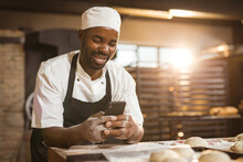 Smiling African American Mid Adult Male Baker Using Smart Phone While Standing In Bakery