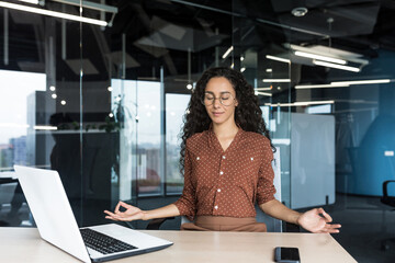 Young beautiful Hispanic web developer working inside office building, woman resting meditating at workplace with eyes closed, female worker using laptop to write code.